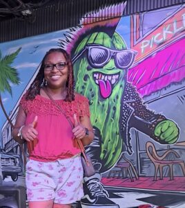 nottypickle Jasmine the Not Typickle owner smiling, standing next to a mural of a cool pickle personified.